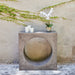 Gray square concrete outdoor console side table with circular cutout, white candles and potted orchid against textured wall with vines