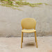 A yellow armless outdoor side chair with a perforated backrest, placed against a plain wall with greenery above