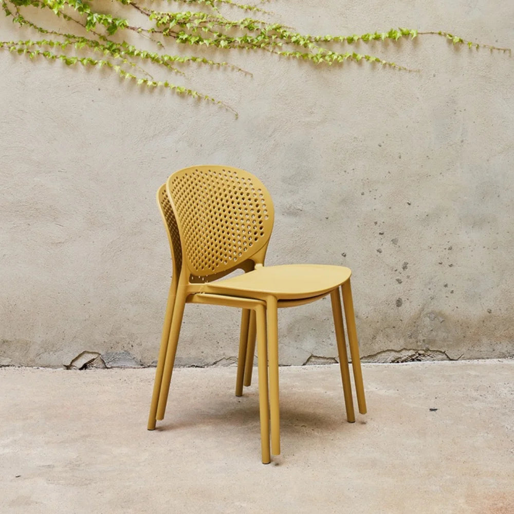 Two stacked yellow outdoor side chairs with perforated backrest against a plain wall with greenery above