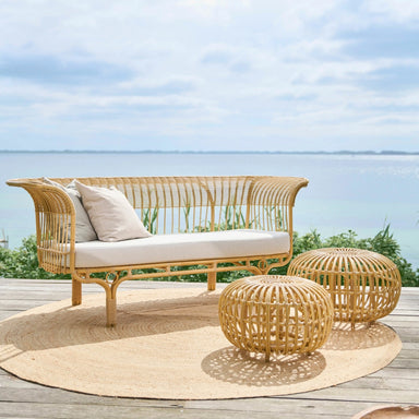 Rectangular wicker outdoor loveseat in natural rattan with curved backrest and beige seat cushions beside round stools.