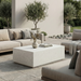 Modern patio with rectangular white concrete outdoor coffee table, beige sofa and chairs, surrounded by greenery