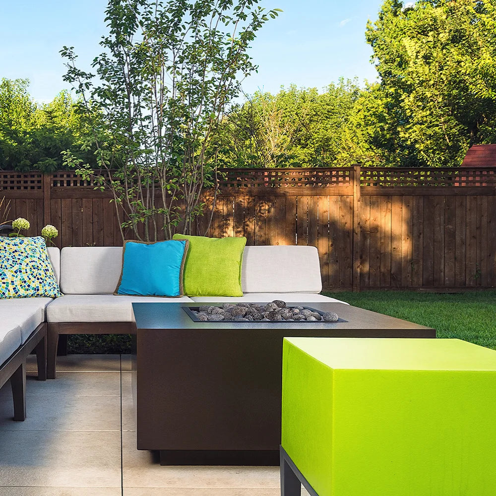 Modern outdoor patio with dark brown square fire pit table, sectional sofa, colorful cushions, and lush green backyard with wooden fence