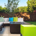 Modern outdoor patio with dark brown square fire pit table, sectional sofa, colorful cushions, and lush green backyard with wooden fence