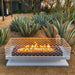 White outdoor rectangular fire pit with geometric cut-out design lit with flames, placed on a soil ground surrounded by agave plants