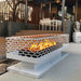 White outdoor rectangular fire pit with geometric cut-out design with glowing flames inside placed on a modern outdoor patio