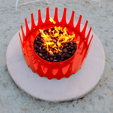 Crown shaped red metal fire pit with burning lava rocks placed on a circular surface over a sandy field
