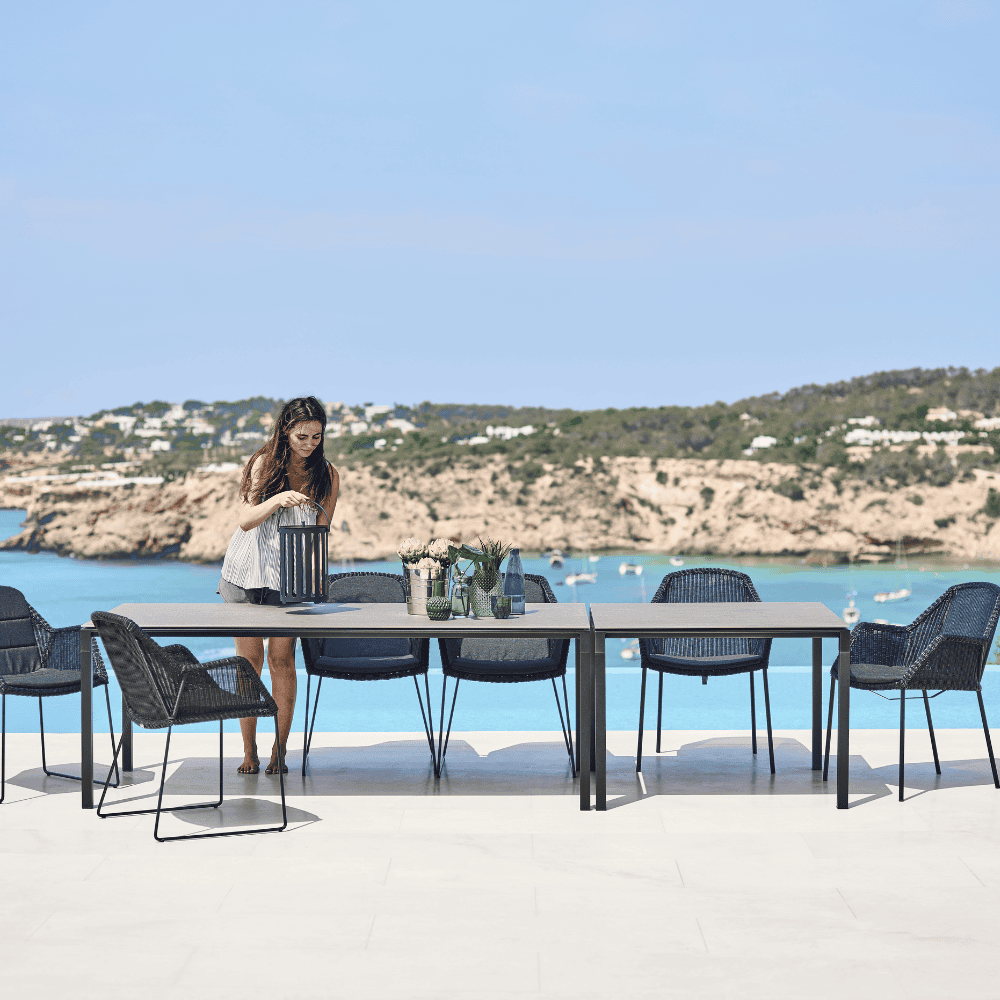 Boxhill's Breeze Dining Weave Chair Black lifestyle image with dining table on rooftop at the seafront with woman standing beside