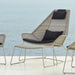 A modern outdoor lounge chair with a woven beige seat and backrest, supported by slim metal legs, featuring two black cushions. A matching ottoman is positioned in front.