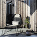 A black woven outdoor lounge chair with a matching footrest and light beige cushions, set on a concrete patio against a weathered wood plank wall with a potted plant nearby.