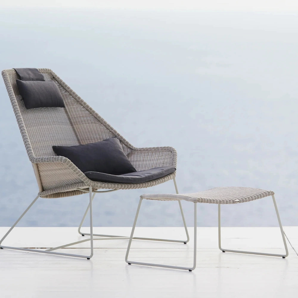 A modern outdoor lounge chair with a matching footstool, both made of woven gray wicker with dark gray cushions, set against a soft, light blue and white background.