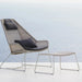 A modern outdoor lounge chair with a matching footstool, both made of woven gray wicker with dark gray cushions, set against a soft, light blue and white background.