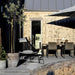 Modern outdoor patio furniture set on a concrete deck in front of a light wood-paneled house. The set includes a black woven lounge chair with a white cushion, a dining table with four matching chairs, and a large patio umbrella casting a shadow.