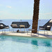 Modern outdoor patio furniture set by a swimming pool with an ocean view, featuring a black woven sofa, two matching lounge chairs with white cushions, a small side table, and a palm tree trunk in the foreground.