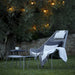 A modern outdoor lounge chair with a matching side table on a grassy lawn, set against a dark olive tree with warm string lights. The lounge chair is gray wicker with gray and white cushions and a draped throw blanket.