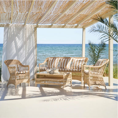 Rectangular natural wicker outdoor sofa with matching chairs and coffee table featuring striped beige and white cushions on a seaside patio.