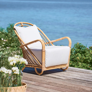 Natural rattan outdoor lounge chair with white cushions on a wooden deck overlooking the ocean.