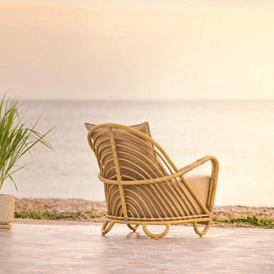 Natural rattan outdoor lounge chair with beige cushions viewed from the back on a seaside patio.