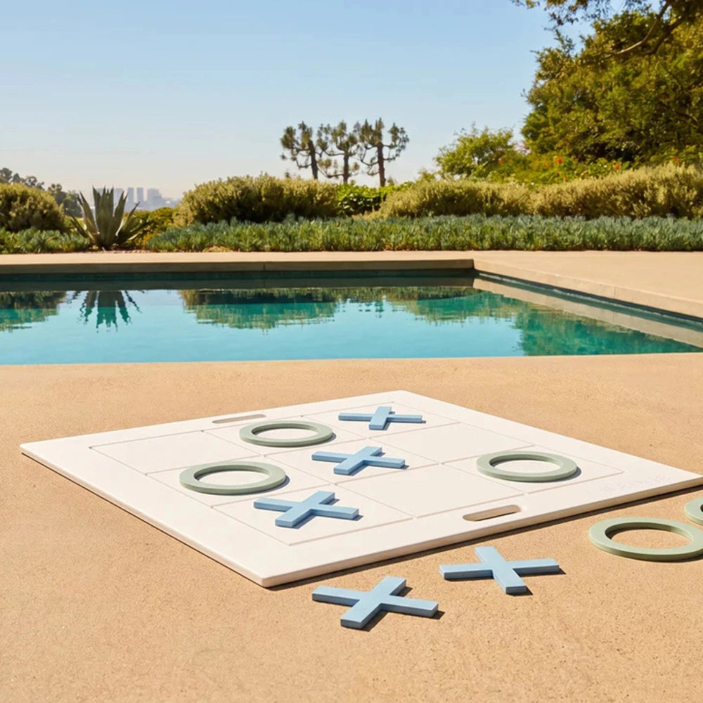 A large white square tic-tac-toe style game board with blue X and O pieces is placed on a sandy poolside deck, with a swimming pool, greenery, and trees in the background.