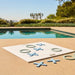 A large white square tic-tac-toe style game board with blue X and O pieces is placed on a sandy poolside deck, with a swimming pool, greenery, and trees in the background.