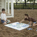 Two children playing a game of tic-tac-toe on a white board outdoors on a mulch-covered playground. One child holds a mint green ring, and the other reaches for a blue cross piece.