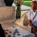 A man in a light blue button-down shirt sits outdoors on a patio, smiling while looking at a checkers game board with blue and green pieces. A patterned throw blanket is draped behind him, with a stone wall, potted plant, and green lawn in the background.