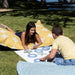 A young woman and a young man are lying on a grassy lawn outdoors, playing with wooden game pieces on a white board. Large yellow and white patterned pillows are behind them, and trees and water are visible in the background.