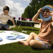Two children outdoors on a grassy lawn playing with light blue circular letter blocks and green letter pieces on a white board. One child holds a blue letter near their face, and the other is seated nearby with more letter pieces.