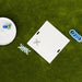 A white square tic-tac-toe game board with a light green cross mark, arranged on a green artificial turf surface. A white plate with blue and green game pieces sits to the left, a stack of white dominoes lies below the board, and a coil of blue rings is to the right.