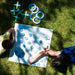 Two children playing a board game outdoors on green grass. The game board is white with blue and green circular tokens, and there are matching blue and green pieces scattered nearby.