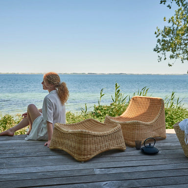 Person sitting beside a natural rattan outdoor lounge chair with matching woven stool on a wooden deck overlooking the ocean.