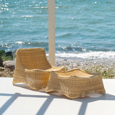 Natural rattan outdoor lounge chair with matching woven stool on a seaside porch overlooking the ocean.