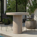 Patio with beige round outdoor bar table and two black bar chairs near glass wall, surrounded by greenery