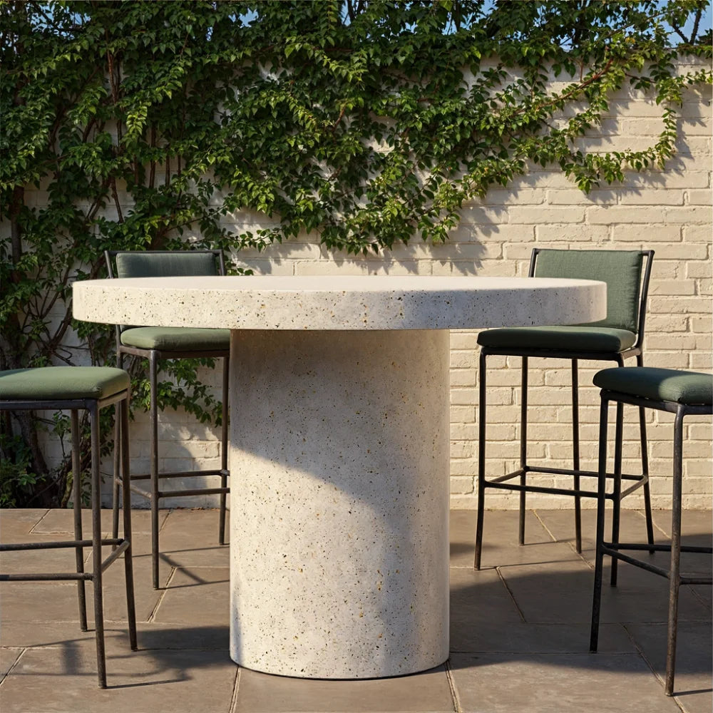 White speckled concrete round outdoor bar table and four green bar chairs on a patio with white brick wall and greenery in the background