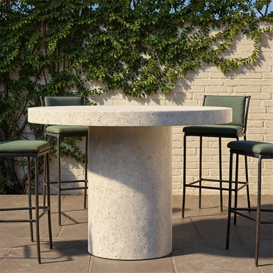 White speckled concrete round outdoor bar table and four green bar chairs on a patio with white brick wall and greenery in the background