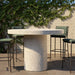 White speckled concrete round outdoor bar table and four green bar chairs on a patio with white brick wall and greenery in the background