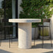 White speckled round concrete outdoor bar table and two black bar chairs on a patio with glass wall and greenery 