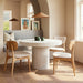 Modern interior setup with white round concrete outdoor dining table, three wooden chairs with white cushions, woven pendant light and gray bench