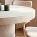 Close-up of white speckled concrete round outdoor dining table and wooden chairs with white cushions in interior space