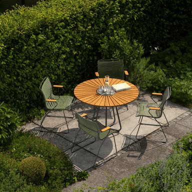 A wooden round outdoor dining table with aluminum base surrounded by green outdoor dining armchairs set on a garden area