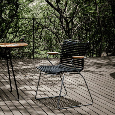 A black outdoor dining chair with bamboo armrest placed on a wooden deck patio surrounded by trees