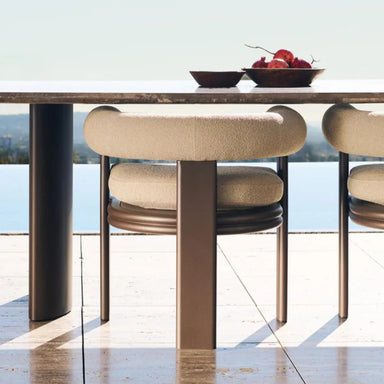 Modern beige outdoor dining chairs with curved barrel-back bolster under a marble-top dining table table with fruit bowls.