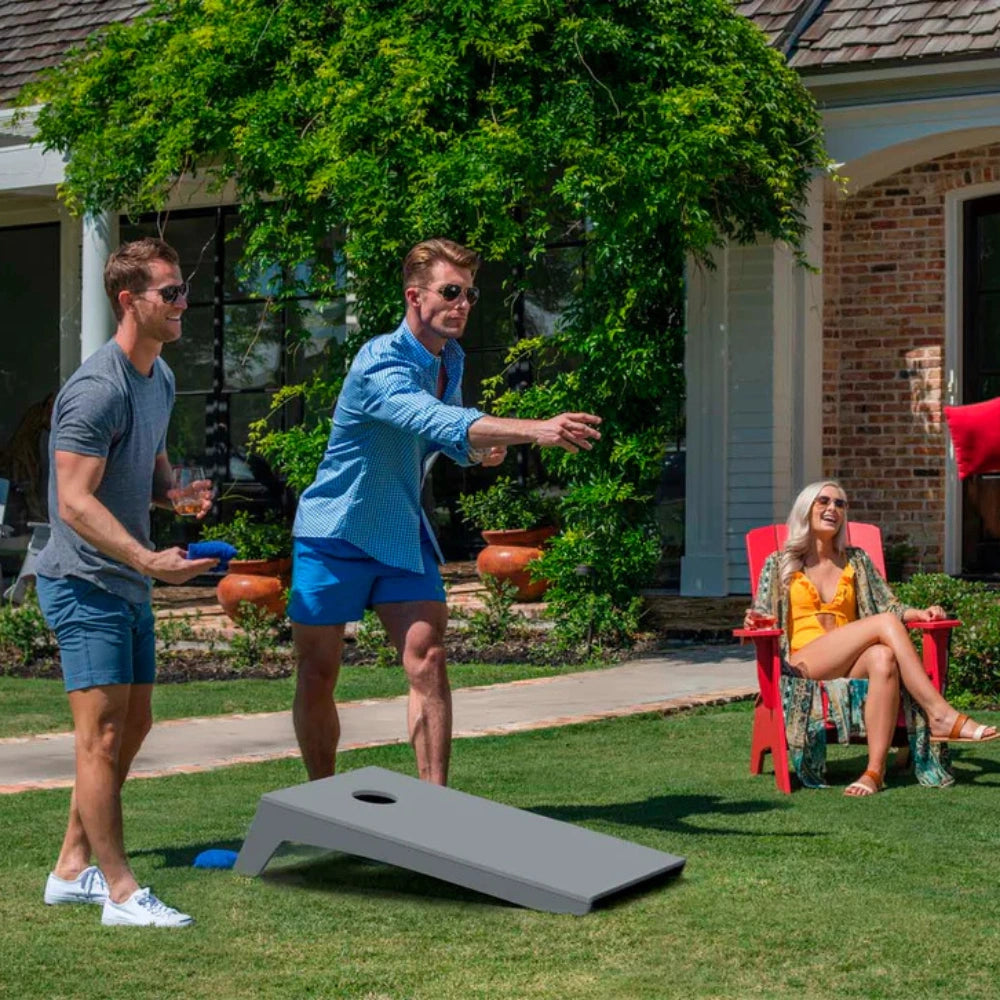 Two men playing cornhole on a green lawn in front of a house, with a woman sitting in a red chair nearby. The cornhole boards are dark gray.
