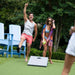 A man and a woman play outdoors on a green lawn with a cornhole board. The man wears a gray tank top, coral swim trunks, and raises his arm in celebration.
