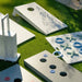 Outdoor cornhole game set on green grass, including white boards with holes, a white scoring/tie-breaker board, and a white tic-tac-toe board with blue and green pieces.