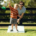 A man and a young boy are outdoors on a white target mat, playing with a green flying disc. The boy is wearing an orange and white striped long-sleeve shirt and gray shorts, while the man wears a light gray t-shirt and sunglasses.