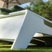 A close-up outdoor shot of a modern white lounge chair on green grass, with dark green cushions and a blurred patio umbrella and trees in the background.