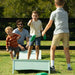 A family playing with green disc golf discs on a grassy lawn. A man and three children are actively engaged in the game, with a white plastic disc golf box in the foreground.
