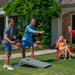 Two men playing cornhole on a green lawn in front of a house, with a woman sitting in a red chair nearby. The cornhole boards are dark gray.