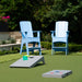 Outdoor backyard scene with two light blue Adirondack chairs on a green lawn, a gray cornhole board in the foreground, and another gray board with bean bags behind it. Trees and hedges are visible in the background.