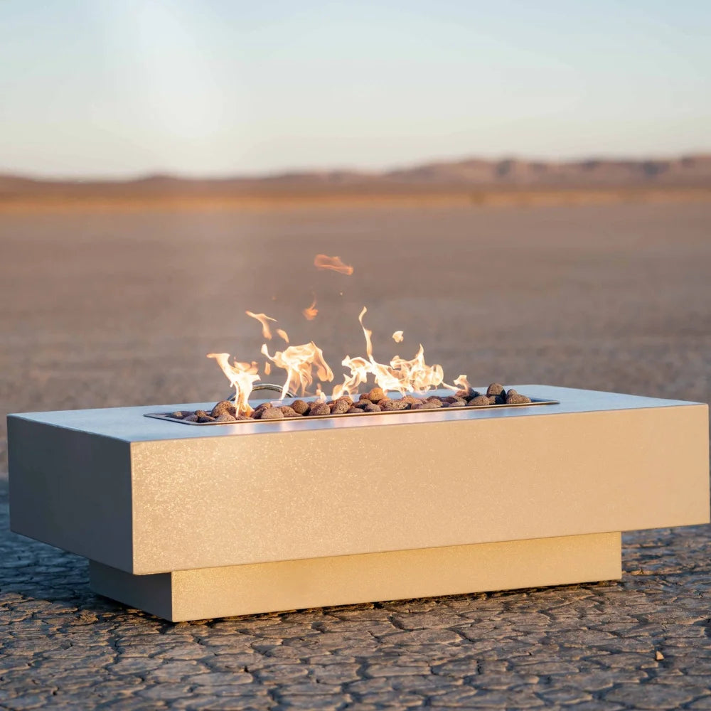 A white rectangular outdoor metal powdered coated fire pit with flames on top placed on a cracked, dry desert ground 